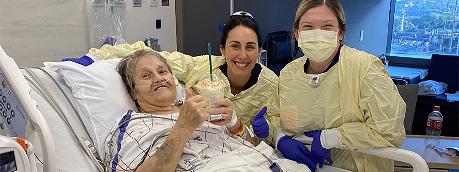 A patient lying in a hospital bed holds up a Starbucks drink she received. Two smiling nurses stand by the bed.