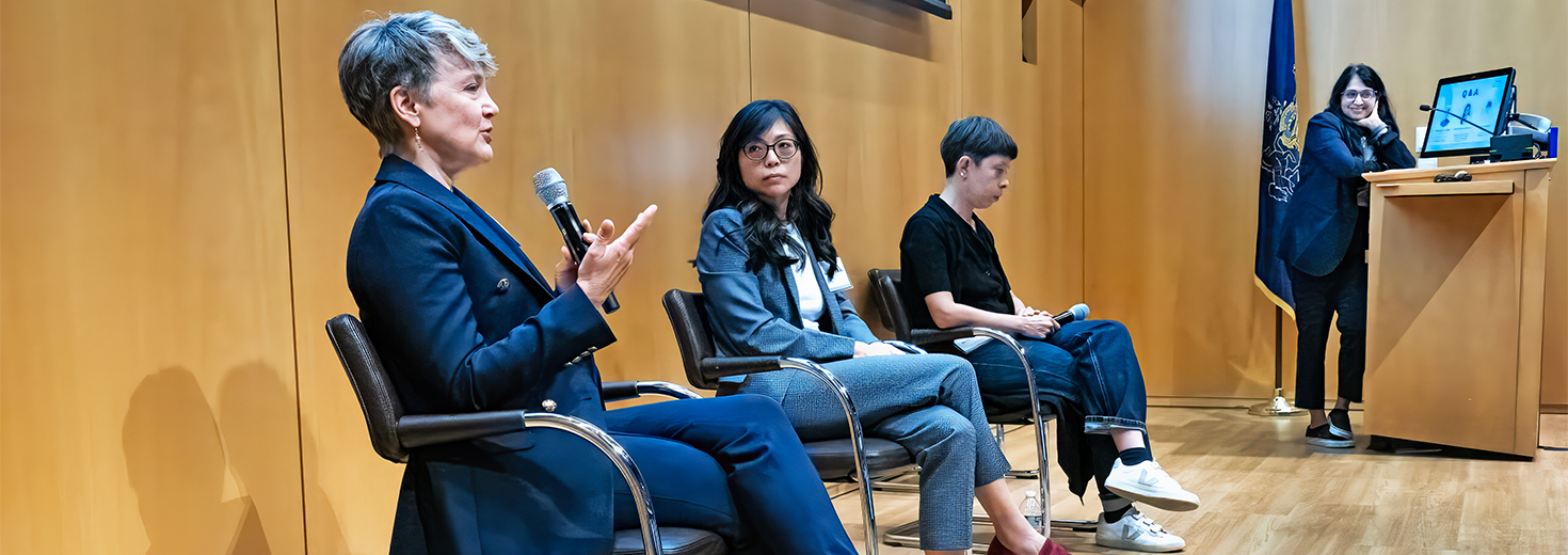 Three speakers sit onstage, with one speaking into a microphone. A moderator stands at a podium in the background.