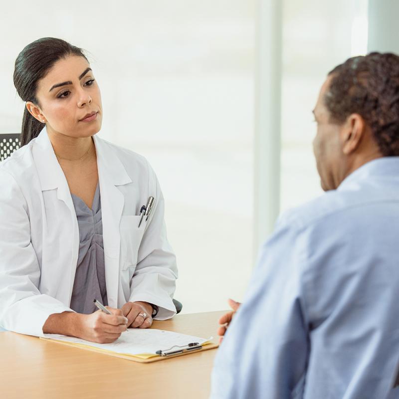 Female doctor listens to man across a desk and holds a pen above a clipboard