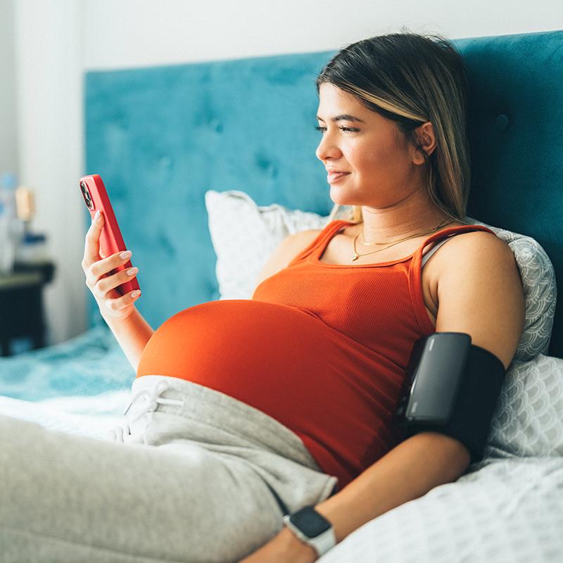Pregnant woman in bed with a blood pressure cuff on one arm and holding a cell phone